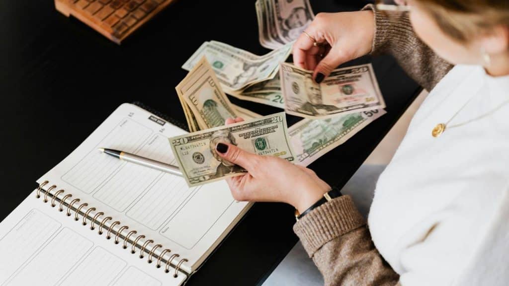 A person counting cash next to an open planner on a desk.