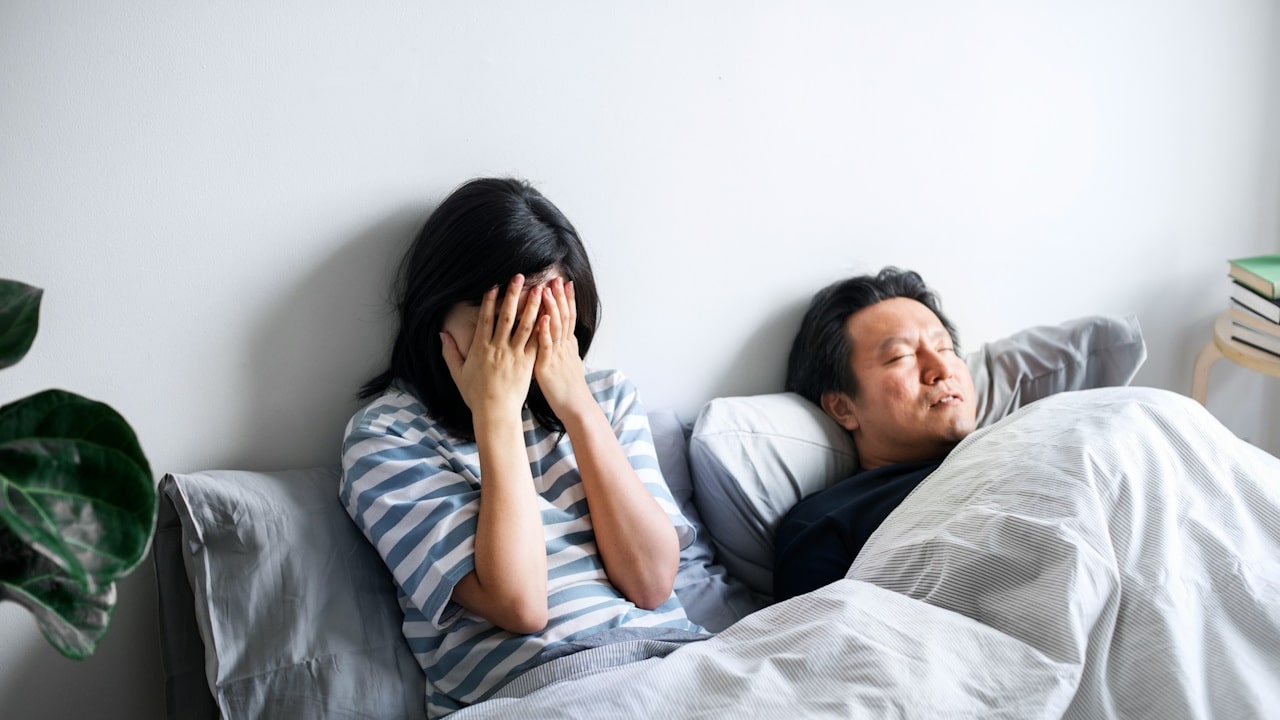 A stressed woman with hands on face sitting in bed with a man sleeping beside her.