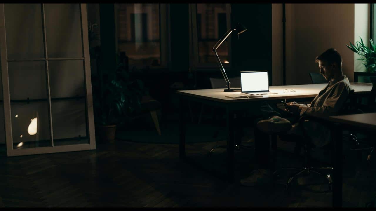 A man sitting at a desk at night, illuminated by a desk lamp and laptop screen.