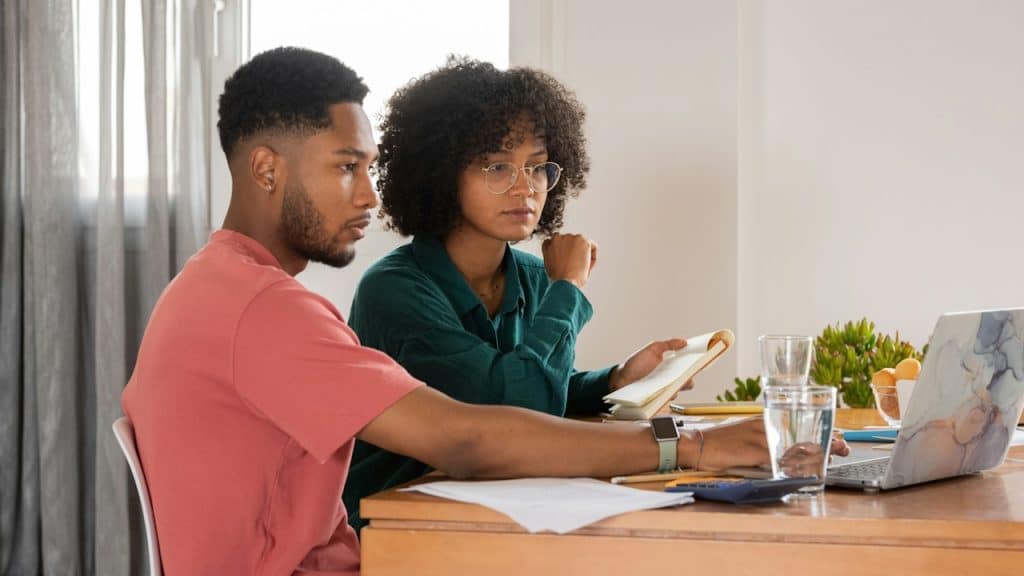 A couple sitting together at a table and looking at a laptop while the woman holds a writing pad in her hand.