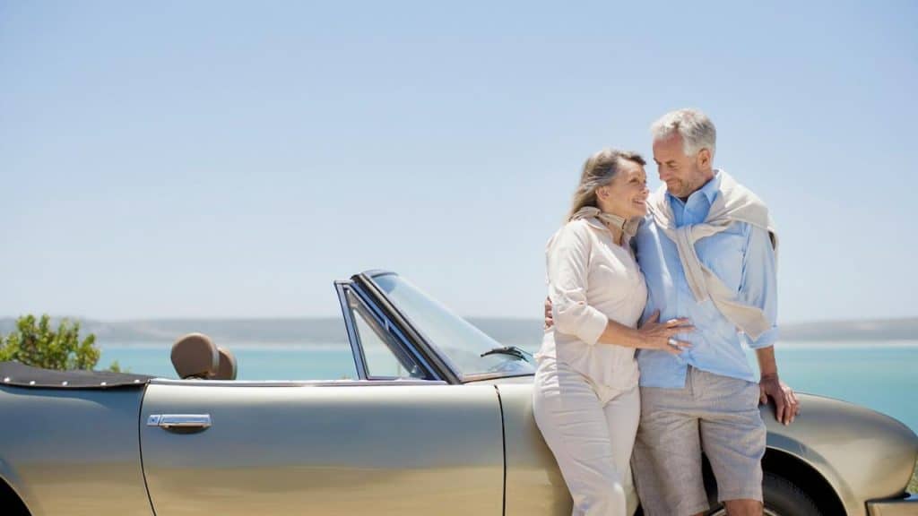 A mature couple hugging and leaning on their convertible with the ocean in the background.