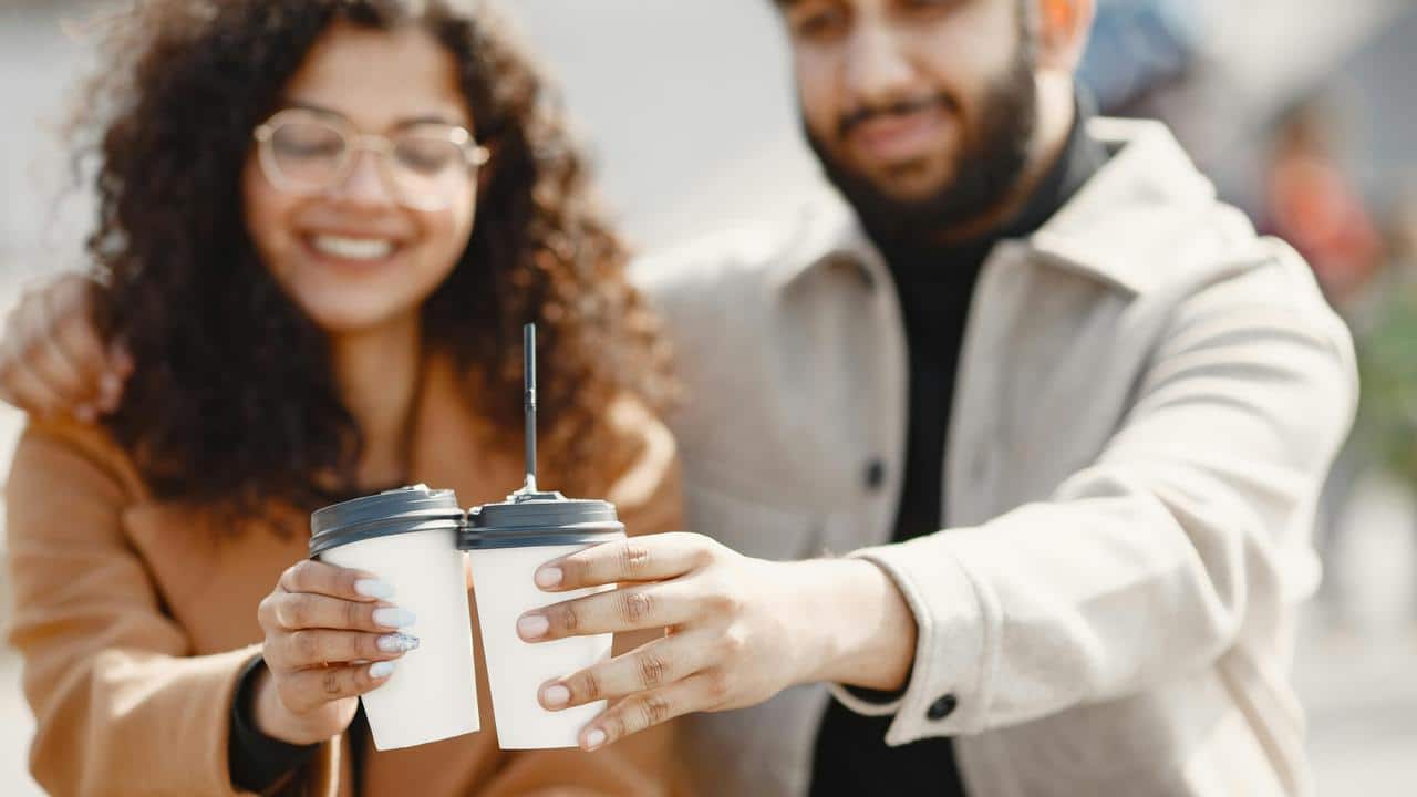 A smiling couple clinking takeaway coffee cups outdoors.
