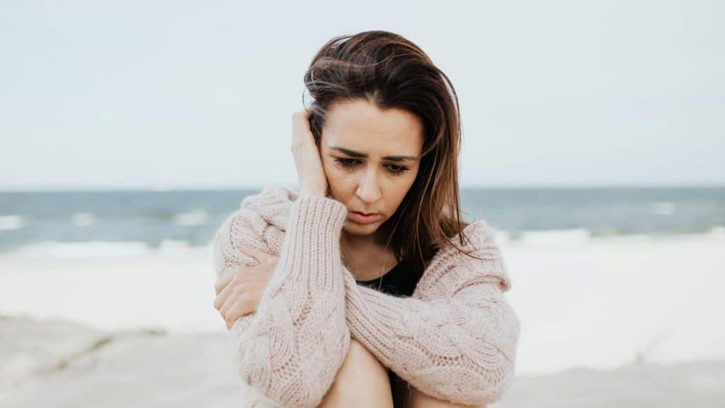 A woman sitting on a beach, looking worried and holding herself.