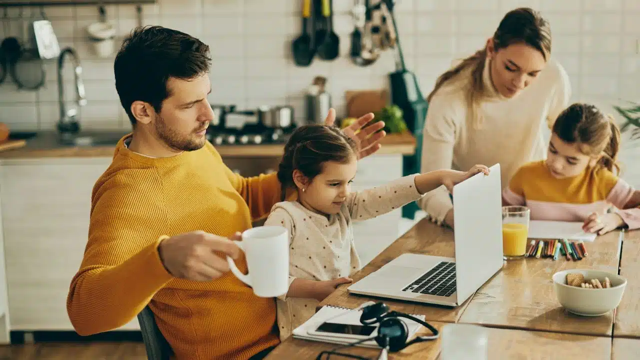 A family of four sits at a wooden table with a laptop, notebooks, and snacks.