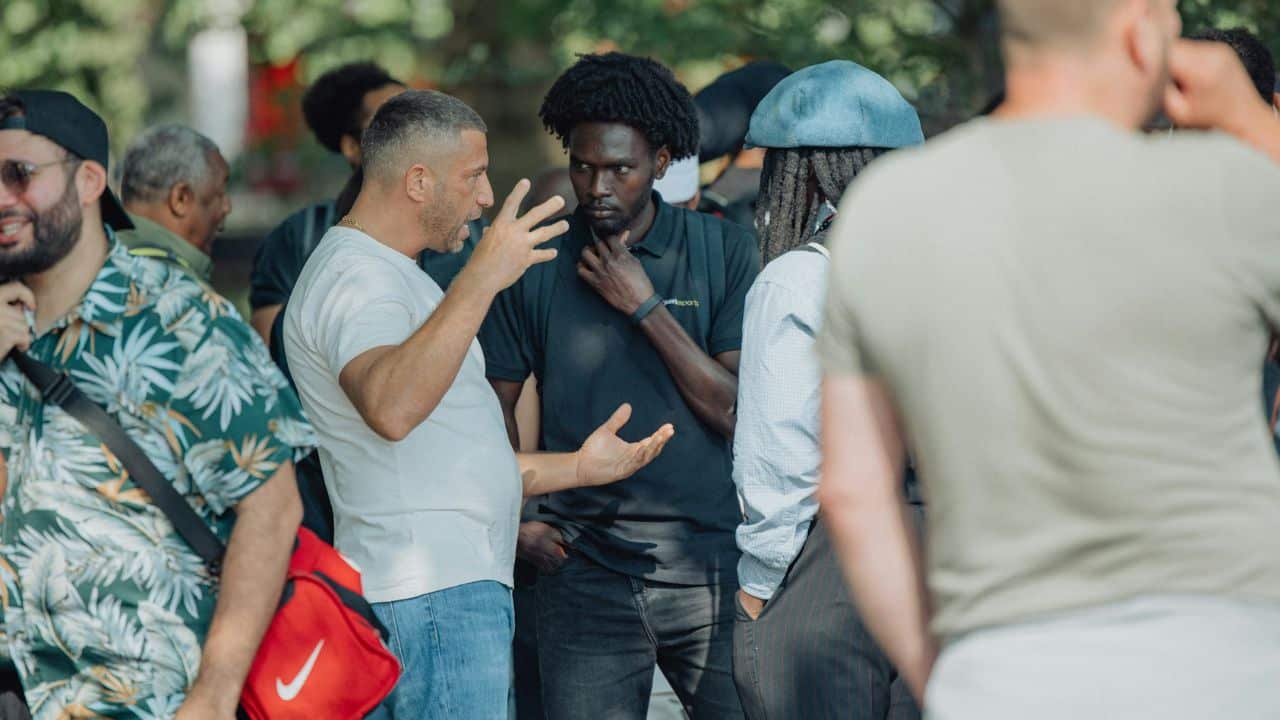A man in a white t-shirt gestures while talking to a group of people outdoors.