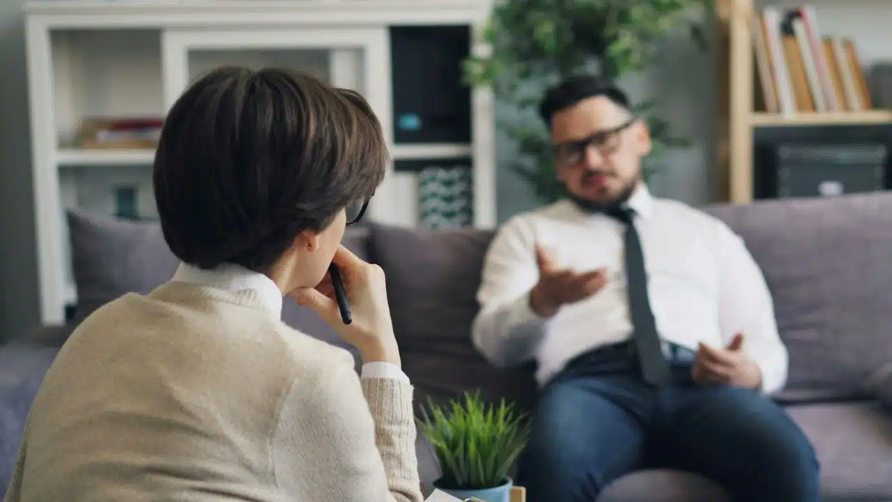 A woman watches a man in a tie gesture while sitting on a purple sofa.
