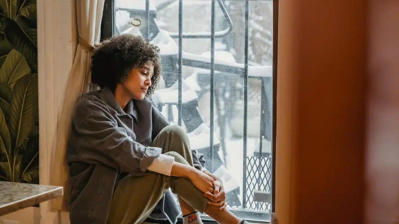 A woman sitting by a window, looking out thoughtfully on a snowy day.
