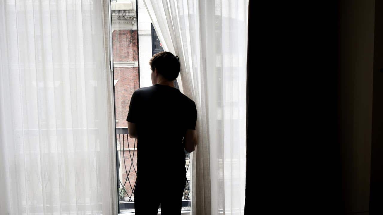 A person standing by a window, looking outside through sheer curtains.