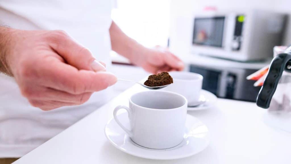 A person spooning coffee grounds into a white cup on a kitchen counter.