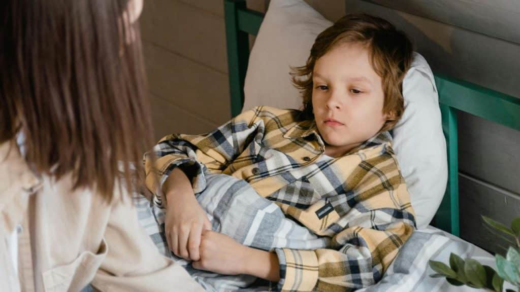 A sick boy lying in bed covered with a blanket while someone sits beside him.