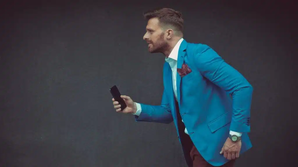 A man in a blue blazer holds a smartphone while leaning forward against a dark background.