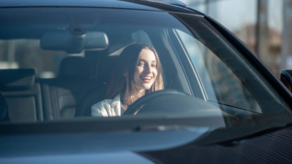 A smiling woman with long hair is seen through a car windshield while sitting inside.