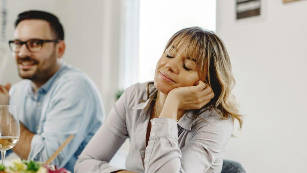 A woman rests her head on her hand with eyes closed near a blurred man.