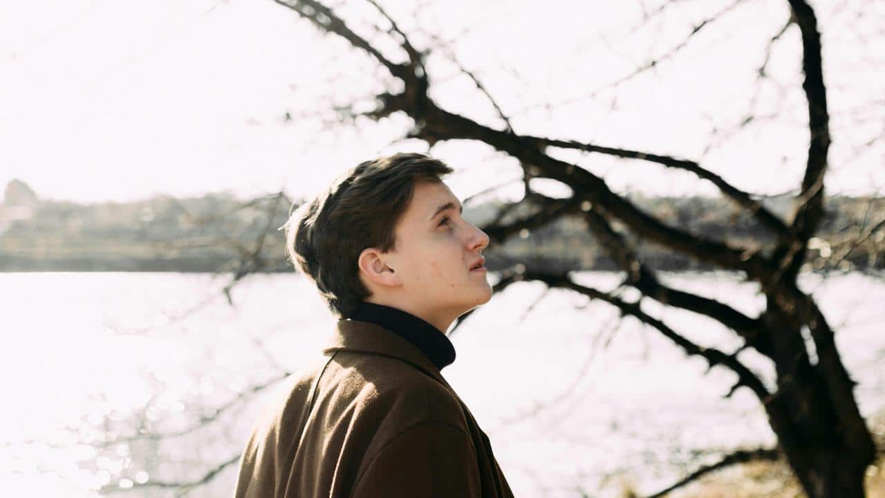 A person outdoors looking upward near a lake and bare trees.