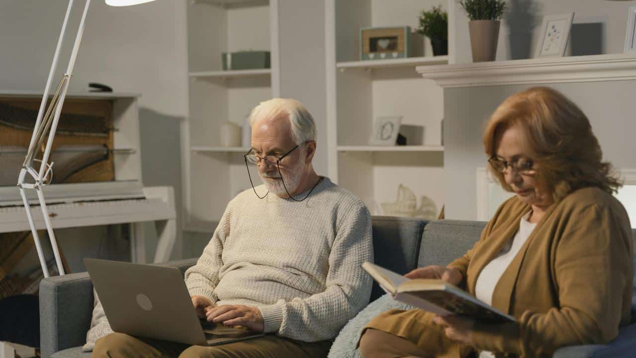 A senior couple sitting on a couch, one using a laptop and the other reading a book.