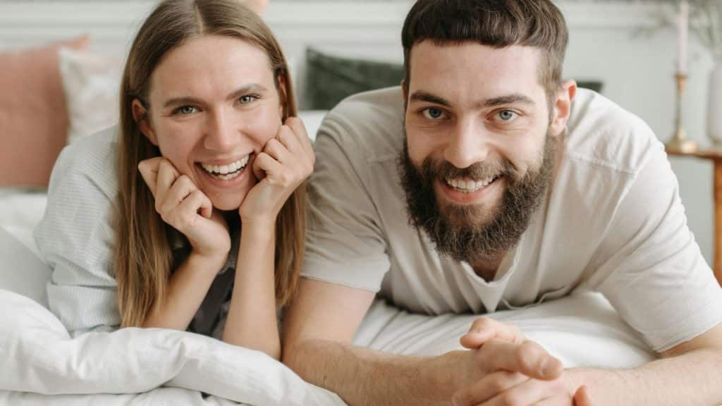 A smiling woman and a bearded man lying down together on a white bed.