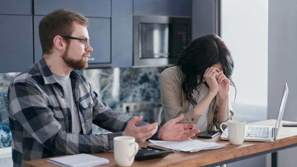 A woman covers her face at a table with a laptop while a man gestures beside her.