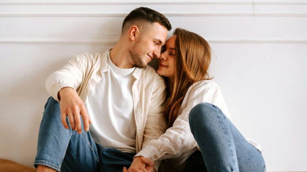 A man and woman sit on the floor, leaning their heads together and smiling.