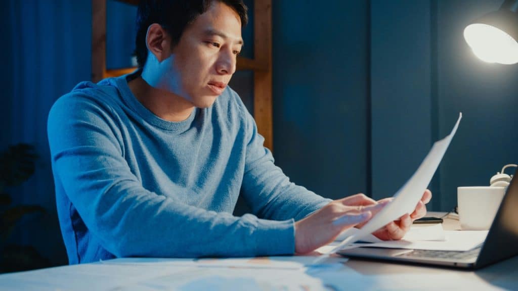 A man in a blue sweater sits at a desk reviewing a paper document.