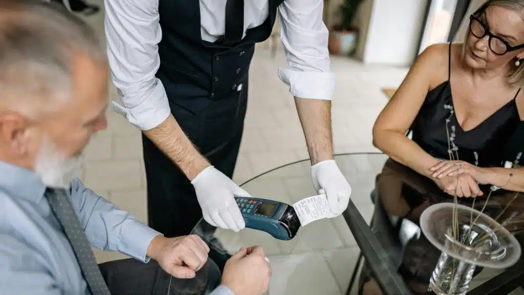 A server in white gloves presents a payment terminal and receipt to a seated couple.