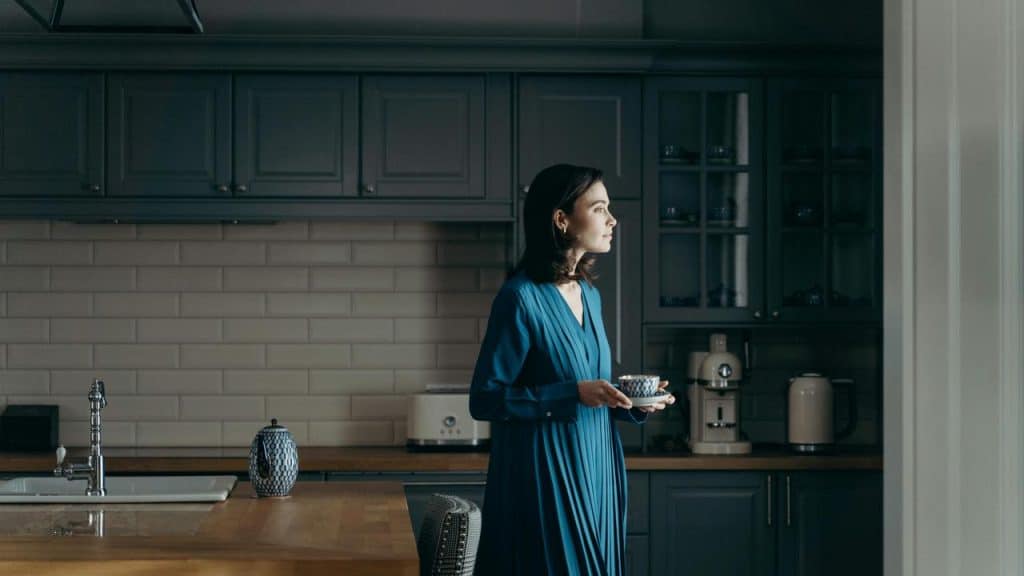 A woman holding a cup of tea while looking out a window in a dim kitchen.