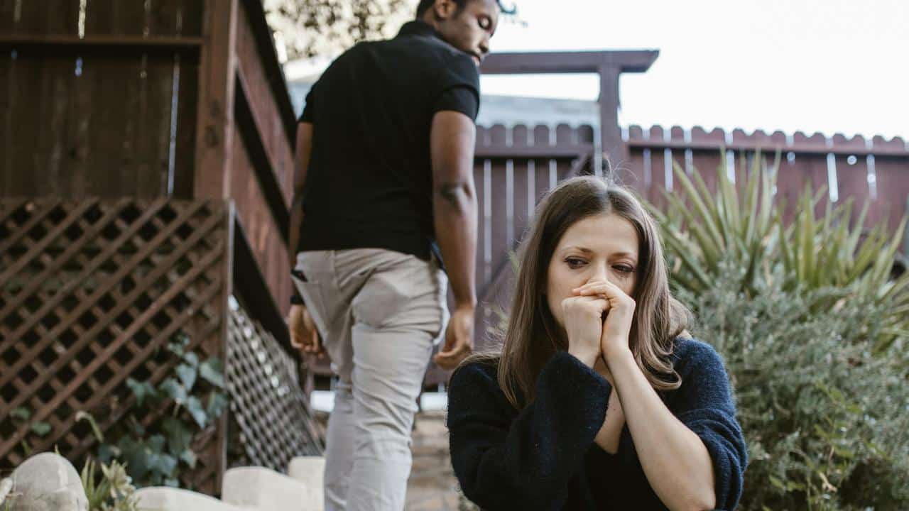 A distressed woman sitting outdoors while a man walks away in the background.