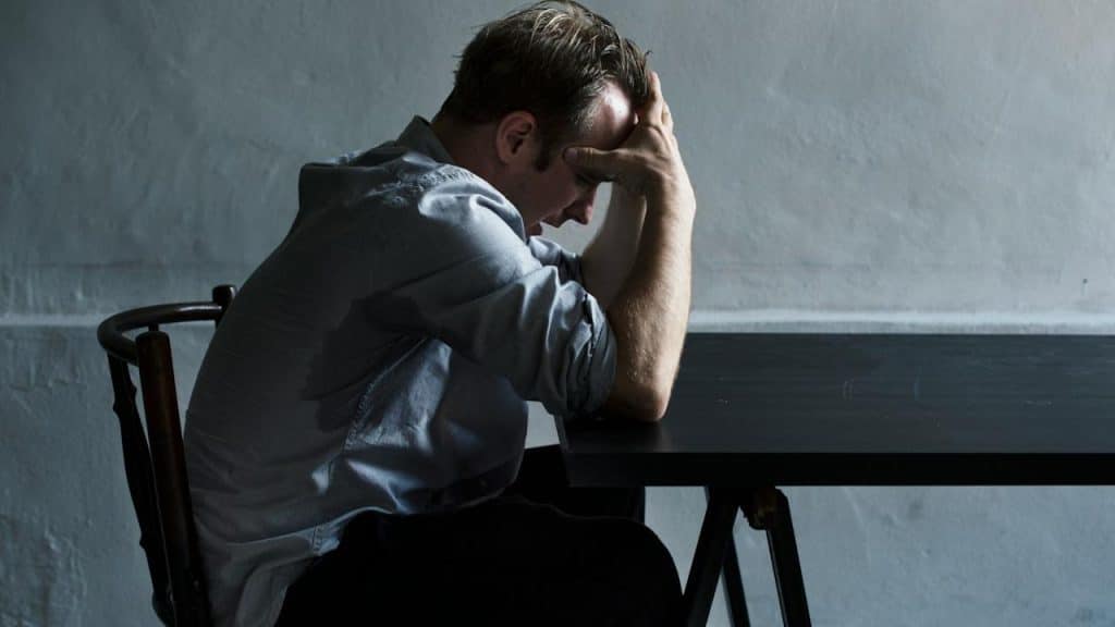 A worried man sitting at a table on a chair and holding his head in his hands.
