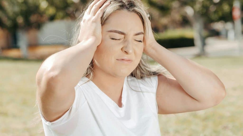A woman outdoors holding her head with eyes closed, appearing stressed or in pain.