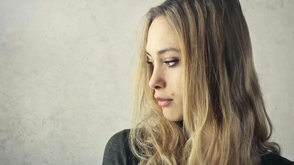 A woman with long blonde hair looking down thoughtfully against a plain background.