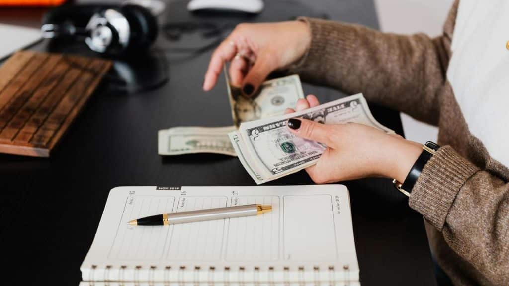 A person counting cash at a desk with a notebook and pen nearby.