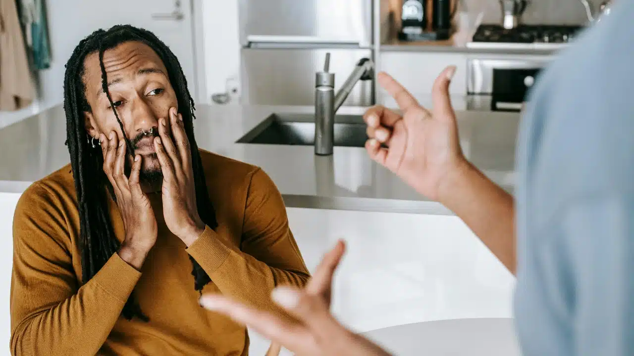 A man with dreadlocks holds his face while another person gestures toward him with hands.