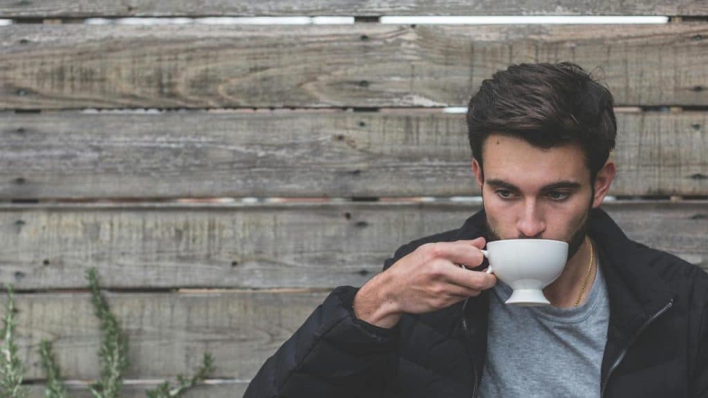 A man drinking from a cup while sitting in front of a wooden wall.