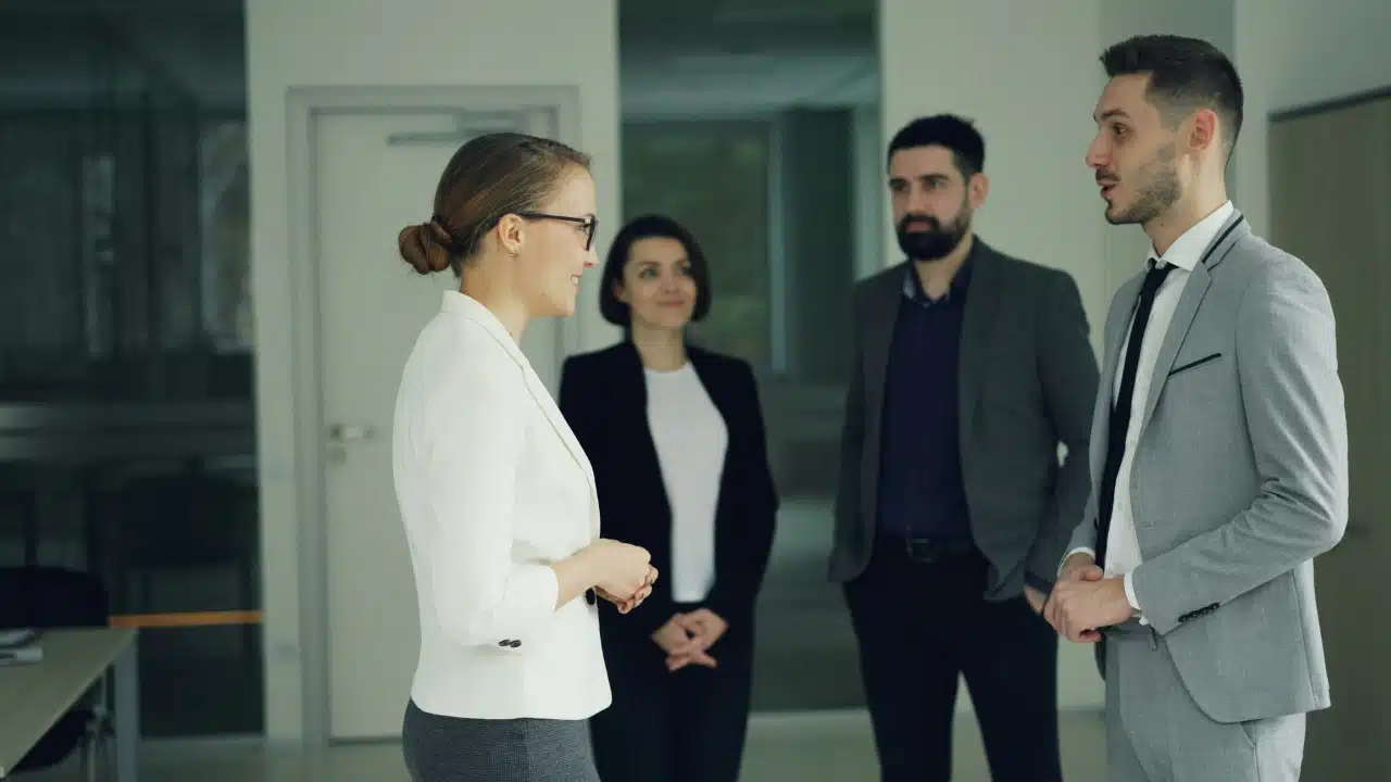 Four people in professional attire stand in an office hallway engaged in a group conversation.