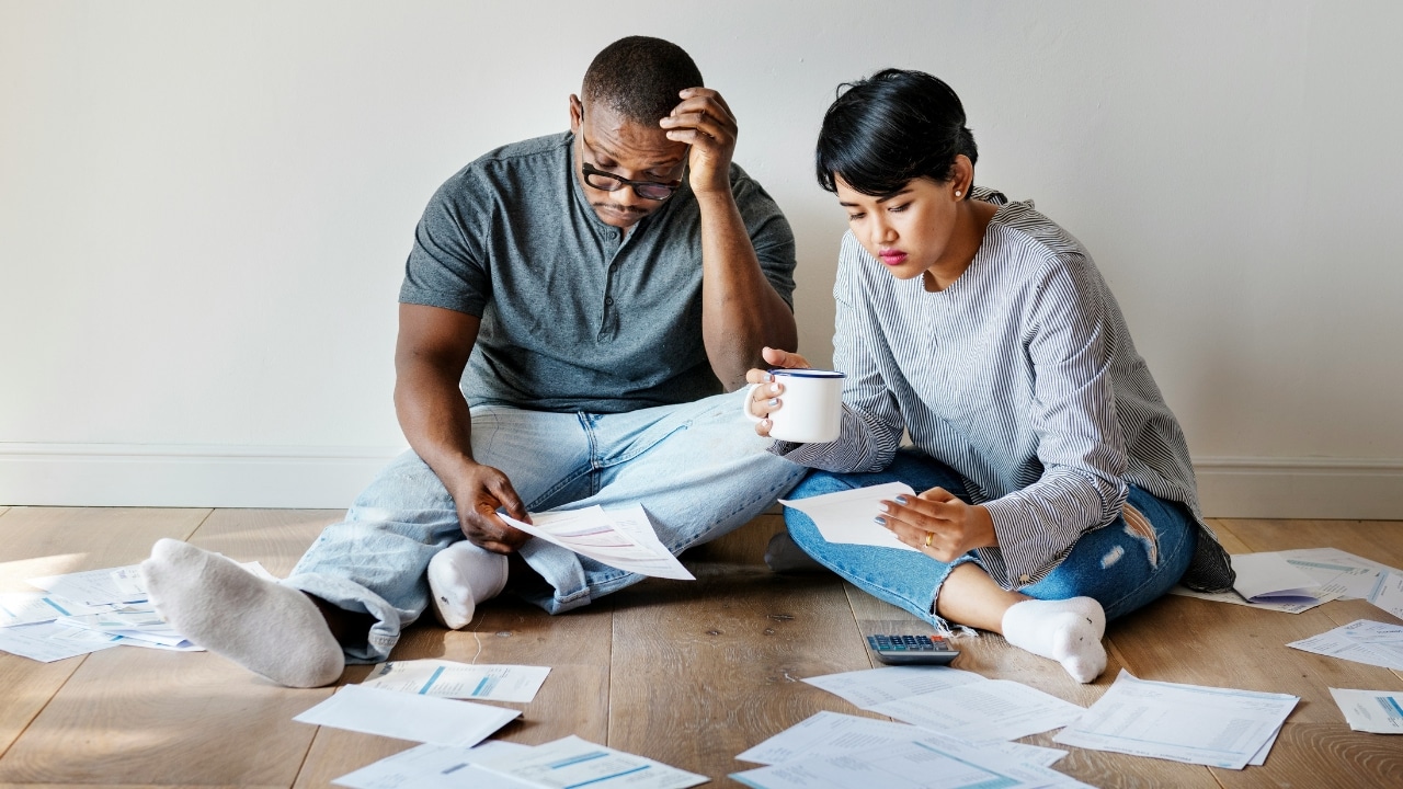 A man and woman sit on the floor surrounded by papers, looking down with concerned expressions.
