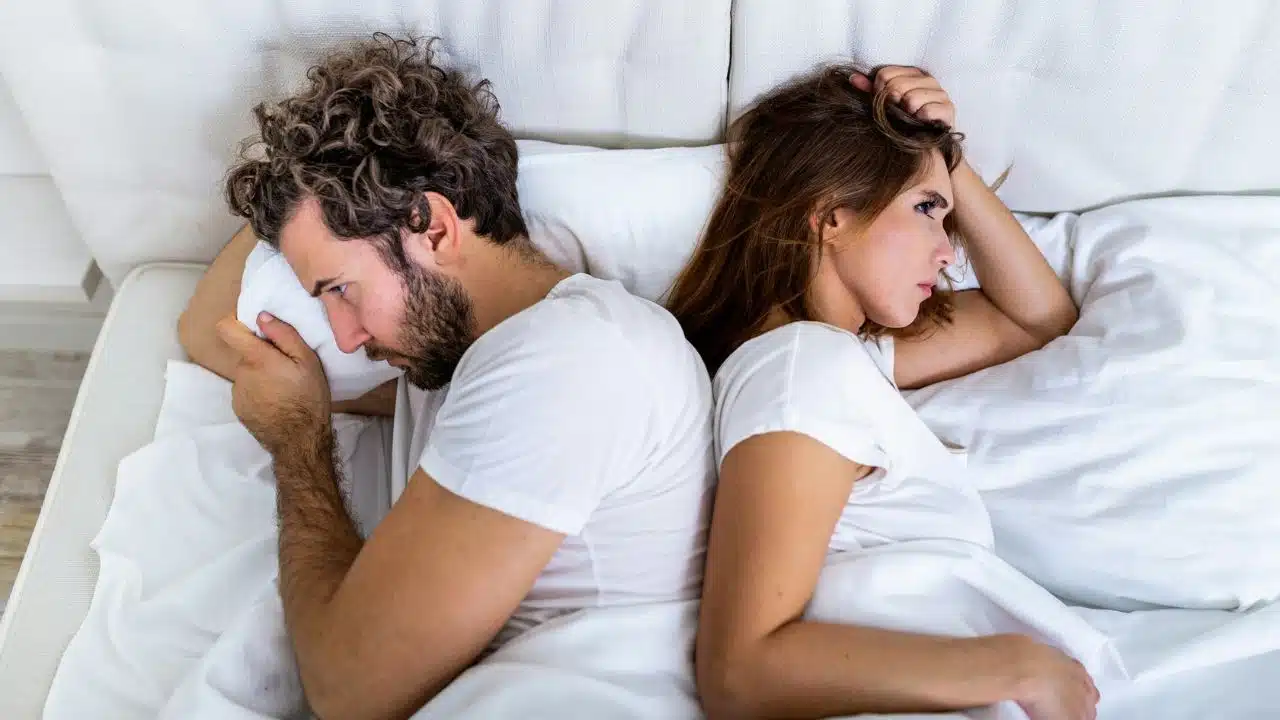 A man and woman lie in bed back to back, looking in opposite directions.
