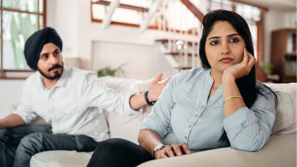 A woman is sitting on a couch with a distant expression while a man in a turban sits behind her, gesturing toward her.