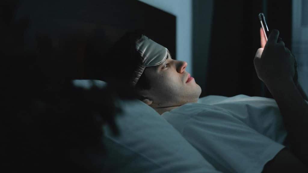 A man lying in bed at night looks at his phone while wearing a sleep mask.