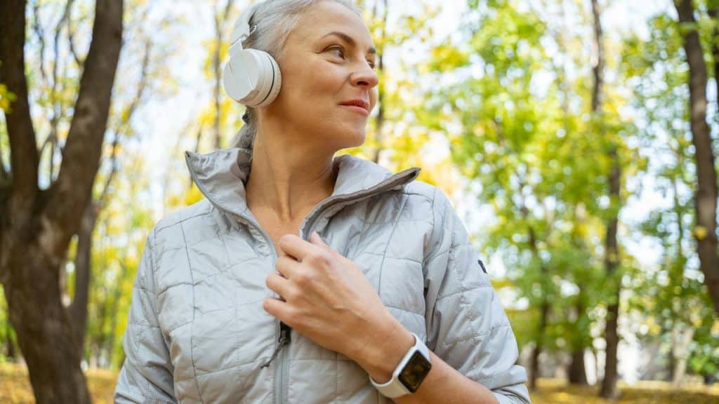 A woman wearing headphones and a light jacket while walking in the park.