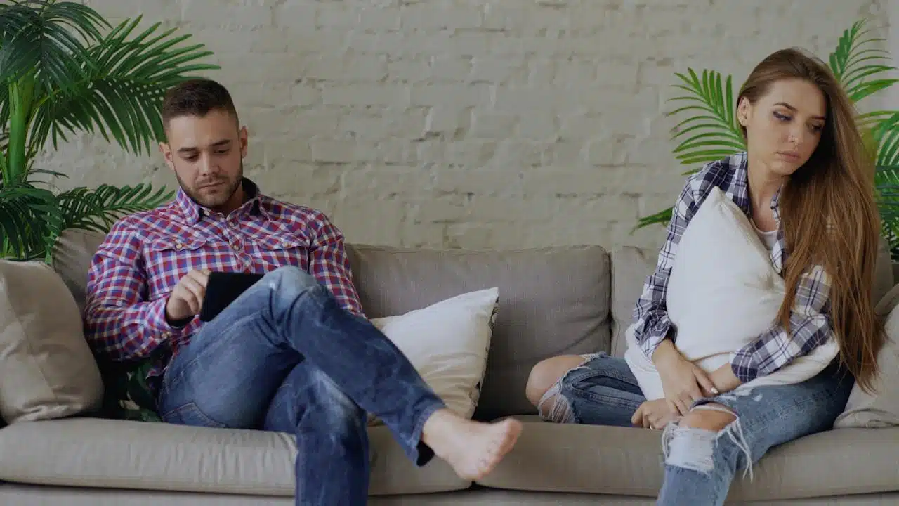 A man is using a tablet while a woman sits on the same couch looking sad, with both ignoring each other.