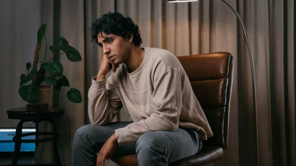 A man with dark curly hair sits in a leather chair with his chin on his hand.