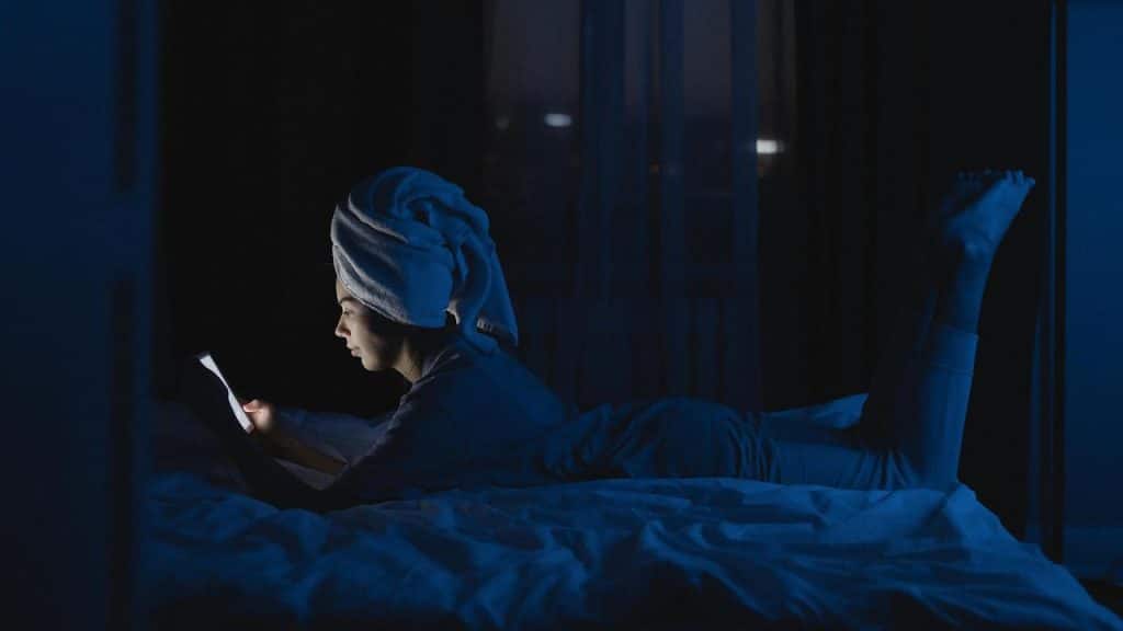 A person lying on a bed at night, reading from a glowing phone.