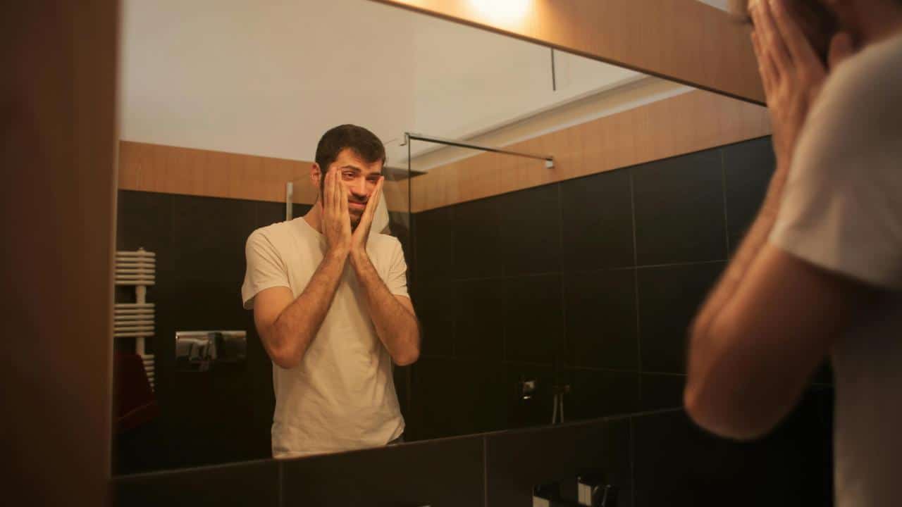 A man touching his face while looking in a bathroom mirror.