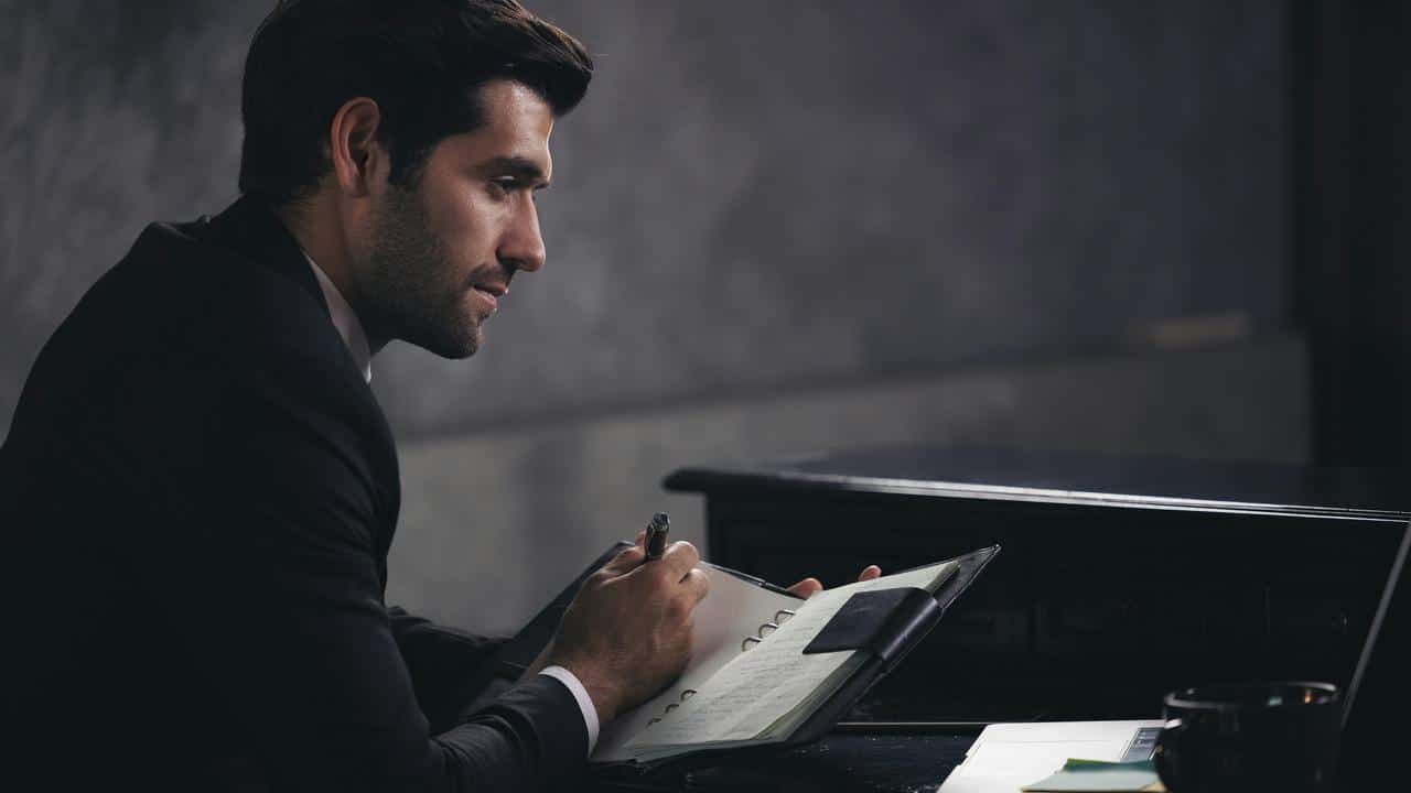 A man in a suit writing in a notebook beside a piano.