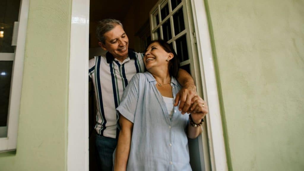 An older man and woman laugh together while standing in a green-painted doorway.