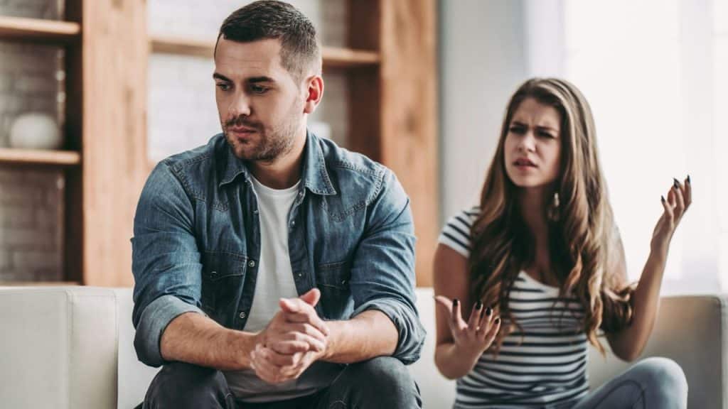A man sits with clasped hands while a woman behind him gestures with frustration.
