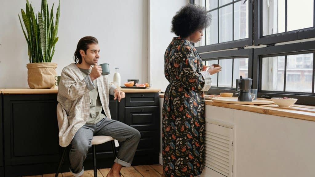 A man drinks from a mug while a woman in a floral robe looks out a window.