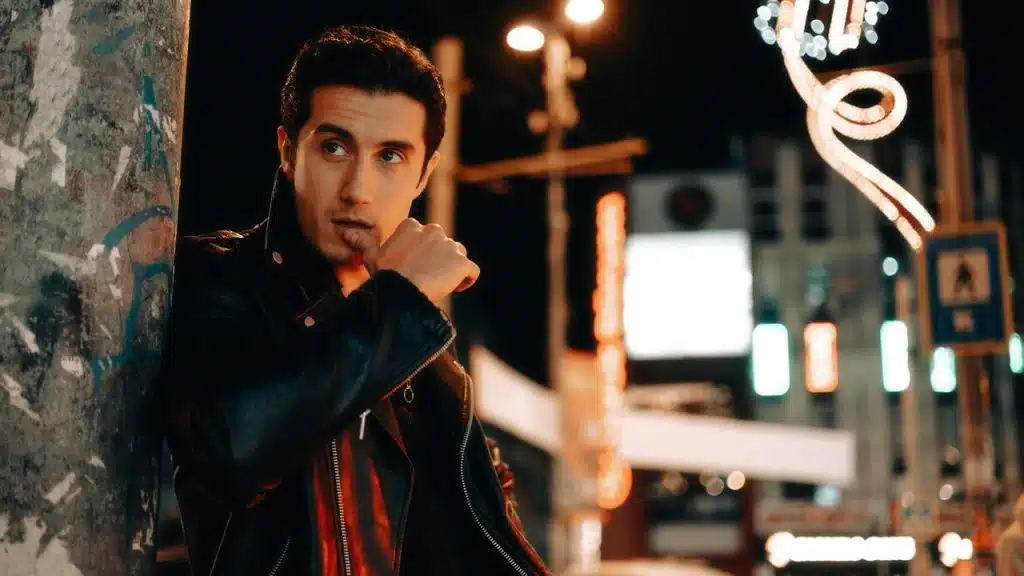 A man in a leather jacket leaning against a wall at night.