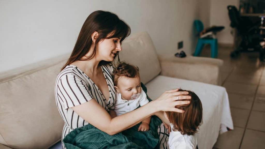 A mother sitting on a couch with a baby while gently touching a child’s head.