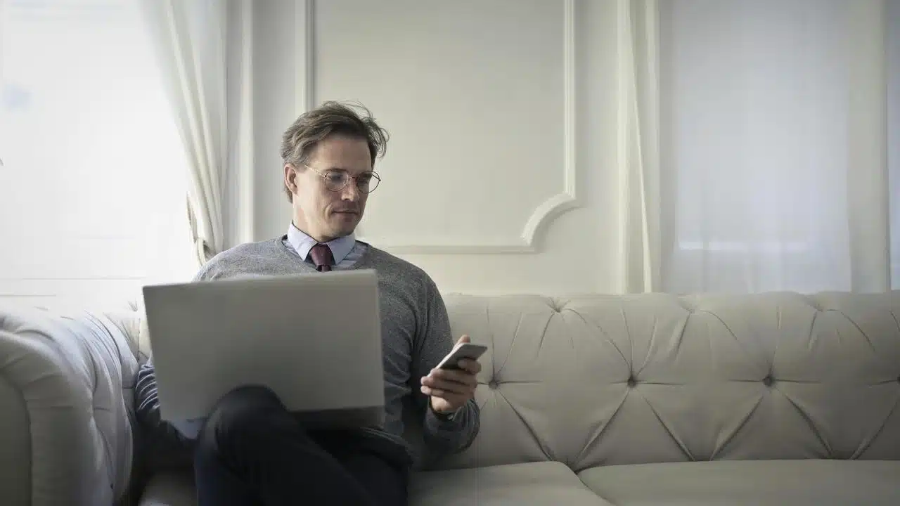 A man sitting on a couch using a laptop and phone.