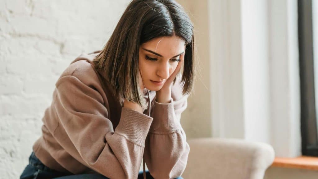 A woman sitting indoors with her hands on her head, looking down thoughtfully.